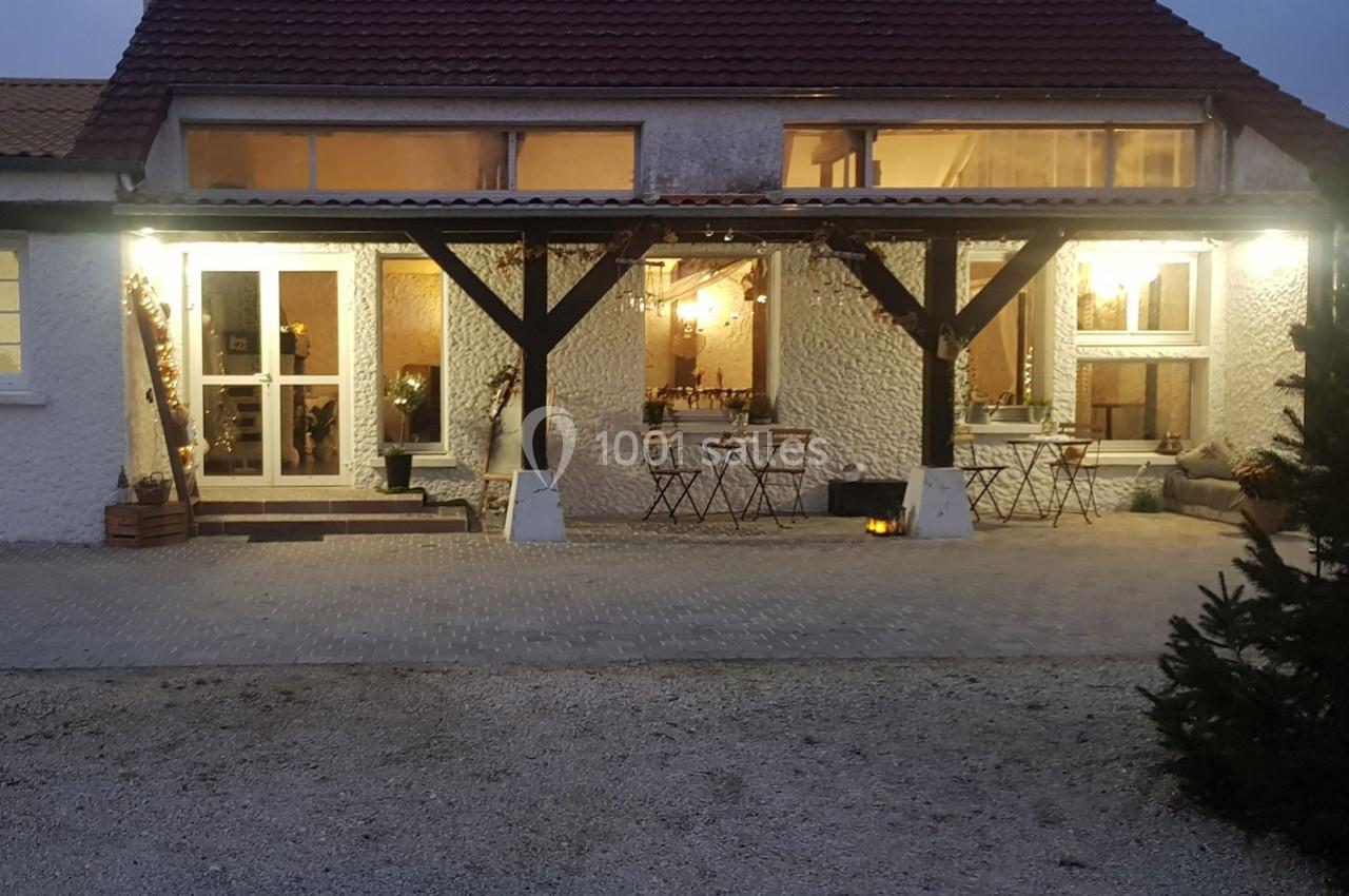 Façade d'une maison avec terrasse éclairée, tables et chaises disposées sous une pergola en soirée.
