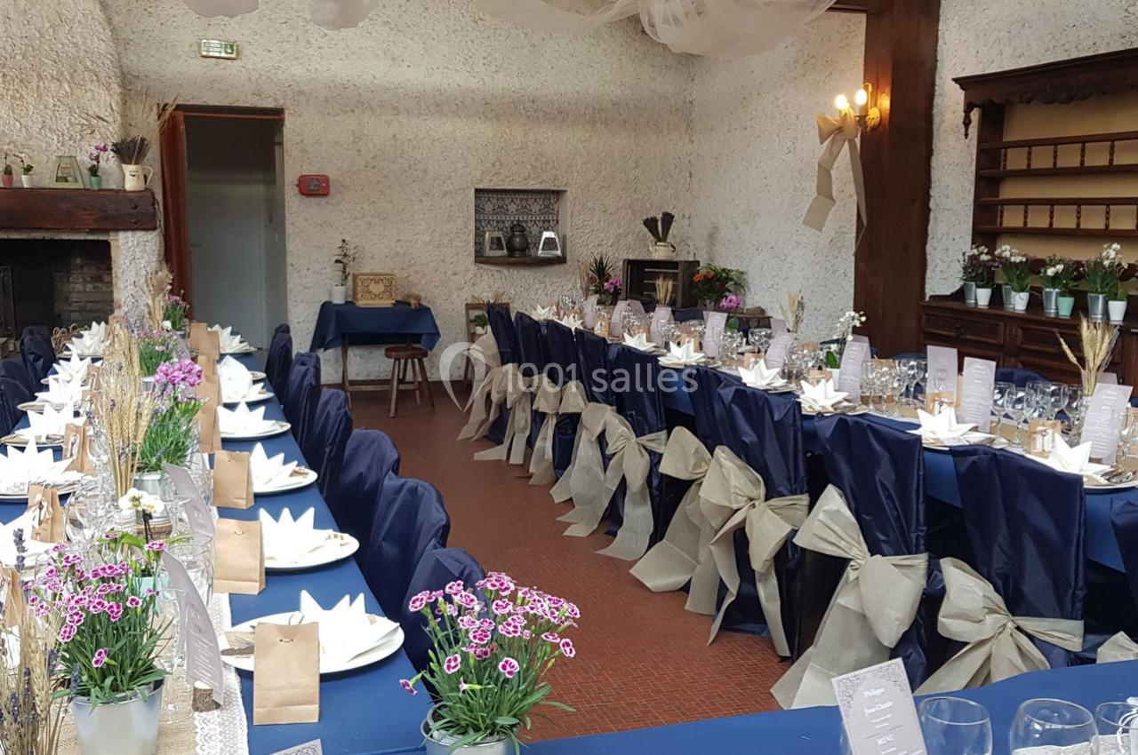 Salle décorée pour un repas festif avec tables alignées, nappes bleues, chaises ornées de nœuds beiges et fleurs en centre…