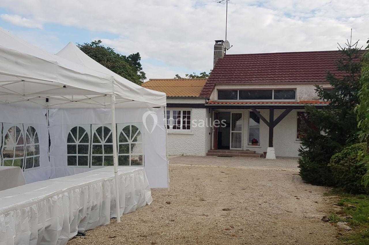 Maison avec toit rouge et auvent, entourée de verdure, avec une tente blanche installée dans la cour gravillonnée.