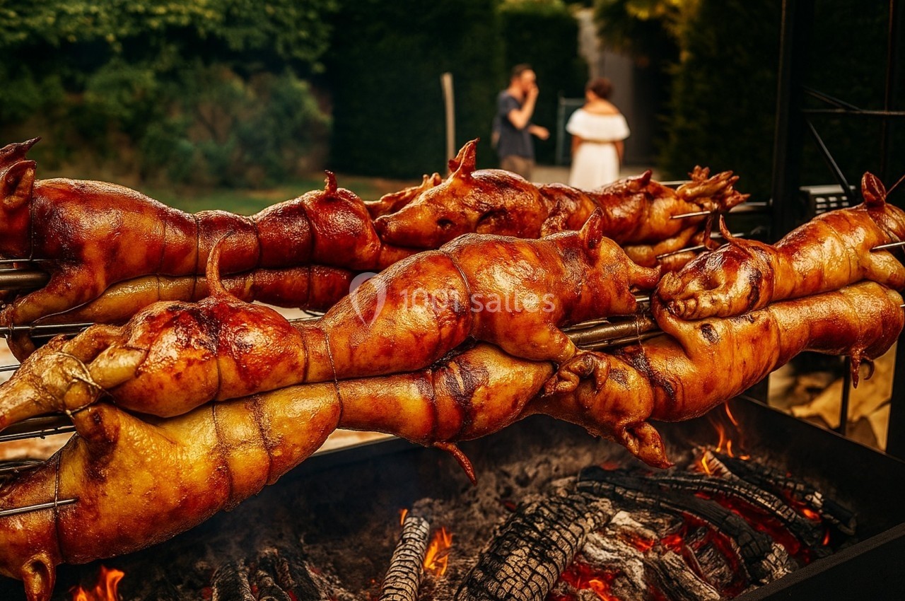 Porcelets rôtis sur des broches au-dessus de braises ardentes, dans un cadre extérieur verdoyant.