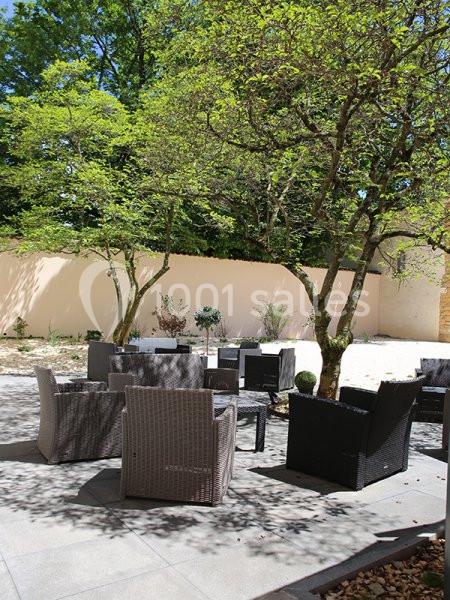 Mobilier de jardin en rotin disposé sur une terrasse ensoleillée, entourée d'arbres et d'un mur clair.