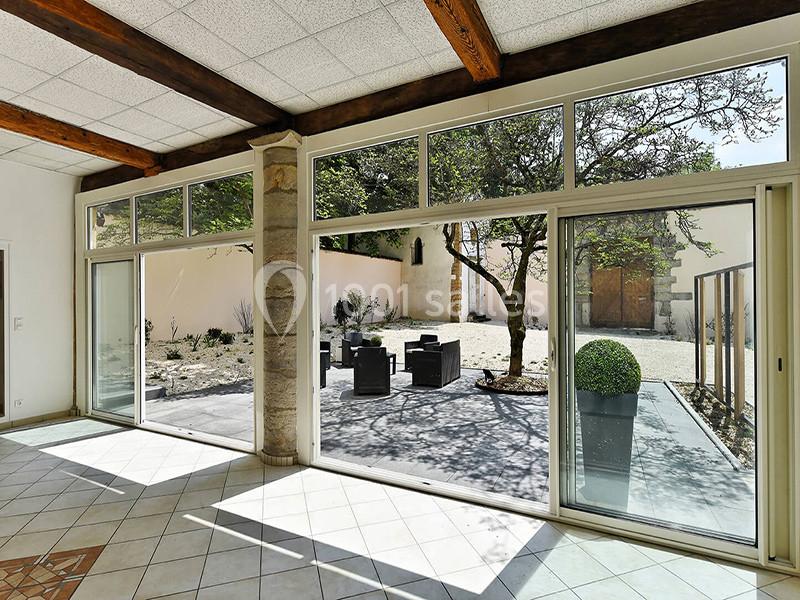 Vue depuis une pièce lumineuse avec baies vitrées donnant sur une terrasse aménagée et un jardin arboré.