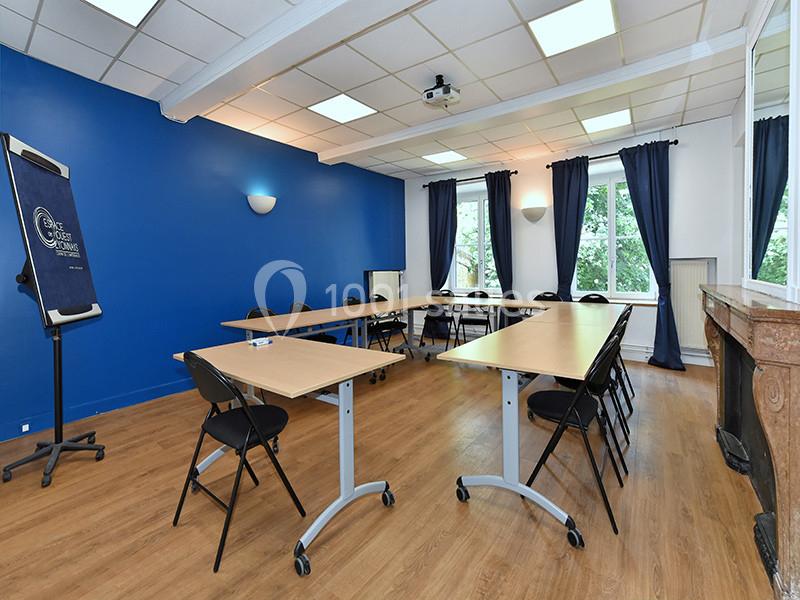 Salle de réunion lumineuse avec tables en U, chaises noires, mur bleu, parquet et fenêtres donnant sur un espace vert.