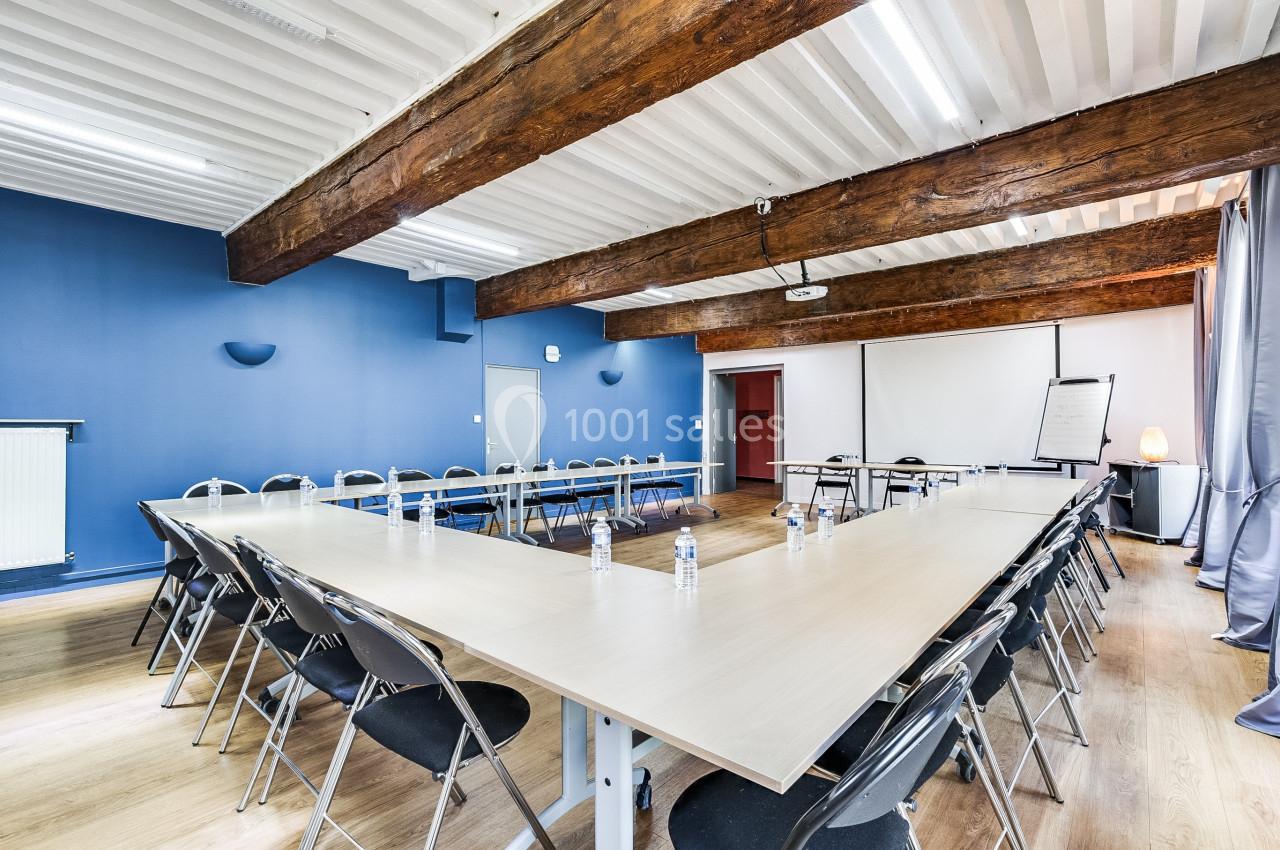 Salle de réunion avec tables en U, chaises pliantes, murs bleus, poutres apparentes et tableau de présentation.