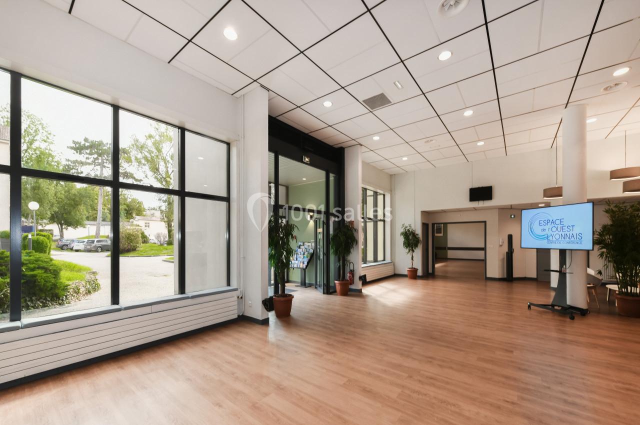 Salle lumineuse avec parquet, grandes baies vitrées donnant sur un espace extérieur verdoyant, et écran d'information.