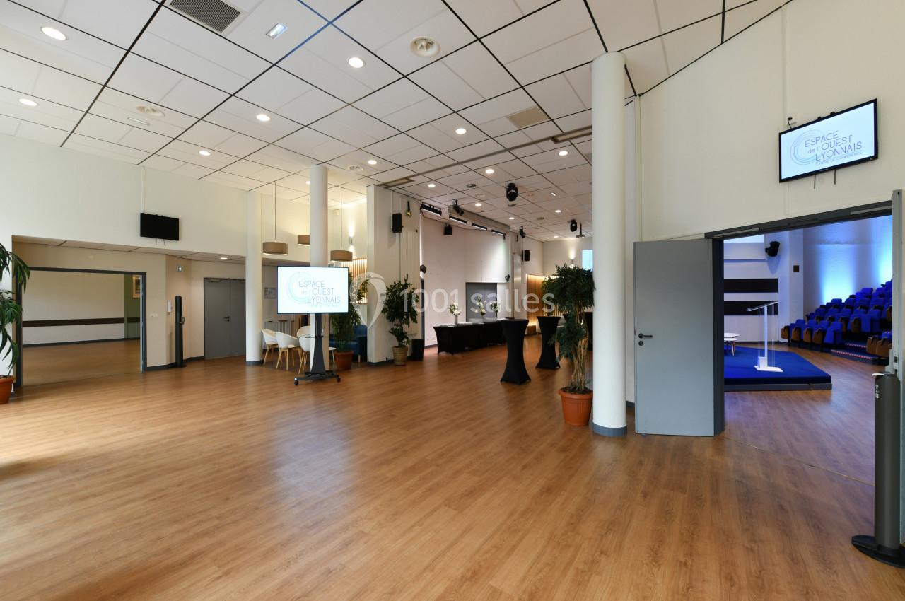 Salle spacieuse avec parquet, écrans, plantes en pot et accès à une salle de conférence avec sièges bleus.
