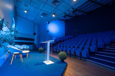 Salle de conférence moderne avec estrade, pupitre transparent, chaises blanches et rangées de sièges vides.