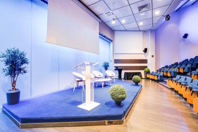 Salle de conférence moderne avec estrade, pupitre transparent, chaises blanches et rangées de sièges vides.