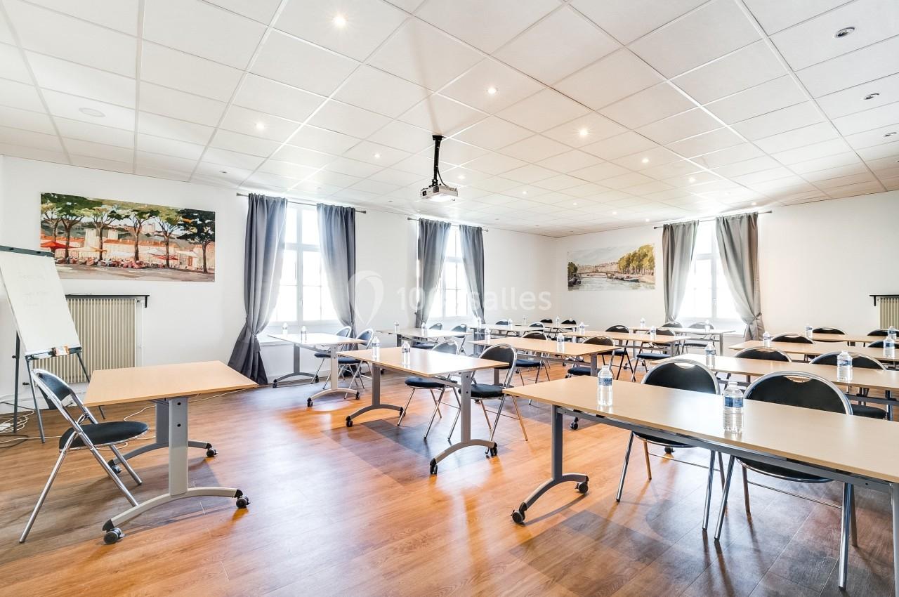 Salle de réunion lumineuse avec tables, chaises, projecteur au plafond et grandes fenêtres avec rideaux.