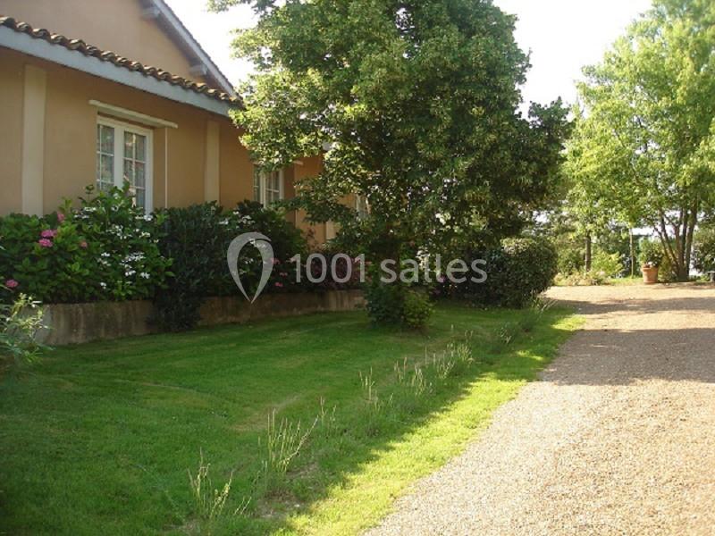 Allée en gravier bordée de pelouse, arbustes et arbres, menant à une maison beige avec des volets blancs.