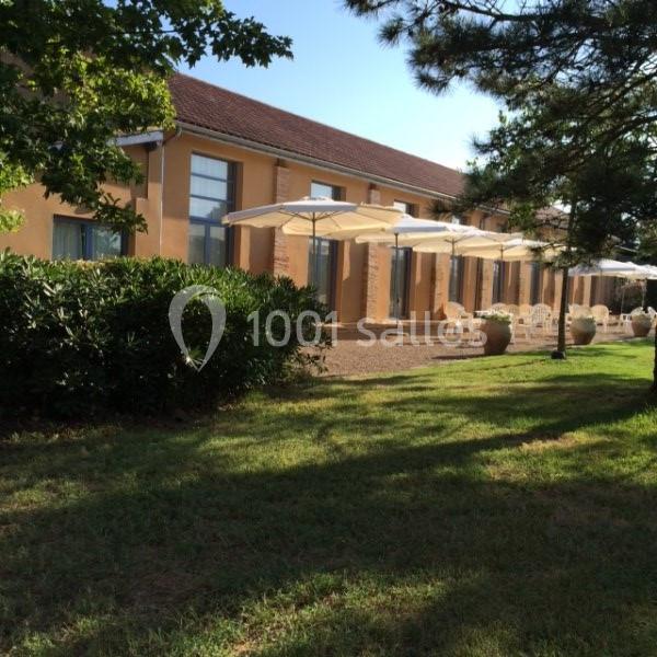 Bâtiment avec façade beige, grandes baies vitrées et terrasses équipées de parasols blancs, entouré de verdure.