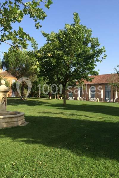 Jardin verdoyant avec pelouse, arbres, fontaine en pierre et bâtiment en arrière-plan sous un ciel dégagé.