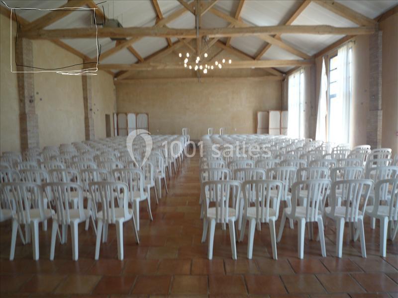 Salle spacieuse avec des rangées de chaises blanches alignées, éclairée par des fenêtres et des lustres suspendus.