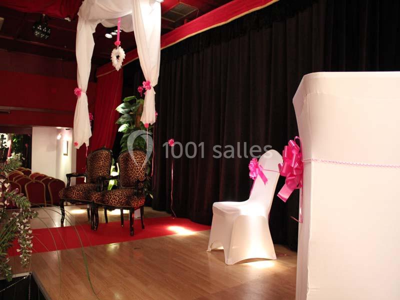 Scène décorée avec des rideaux noirs, fauteuils à motif léopard, chaise blanche ornée d'un ruban rose et éléments floraux.