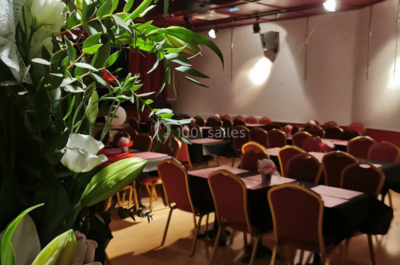 Salle de réception avec des chaises alignées et des tables dressées, vue partielle d'un bouquet de fleurs au premier plan.