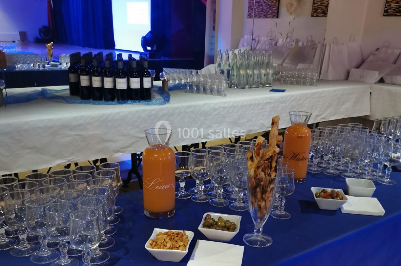 Table dressée avec boissons, verres, carafes de jus, bouteilles de vin, amuse-bouches et décor de salle en arrière-plan.
