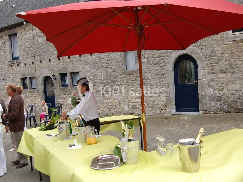 Table dressée avec nappes jaunes, seaux à champagne et verres, sous un grand parasol rouge devant un bâtiment en pierre.