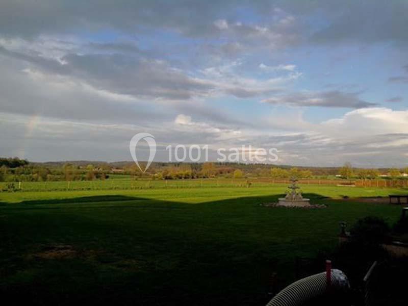 Vue d'un grand jardin verdoyant sous un ciel partiellement nuageux, avec une fontaine au centre et des collines à l'horizon.
