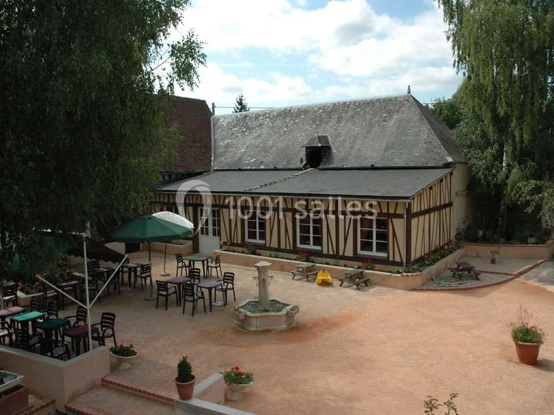 Cour avec une fontaine centrale, entourée de tables et chaises, devant un bâtiment à colombages sous un ciel dégagé.