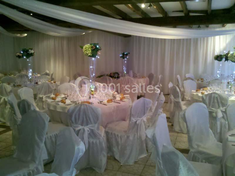 Salle de réception décorée avec des tables rondes, nappes blanches, chaises houssées et centres de table fleuris.