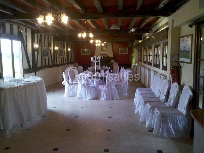 Salle de réception avec chaises blanches couvertes, table dressée au centre et poutres apparentes au plafond.