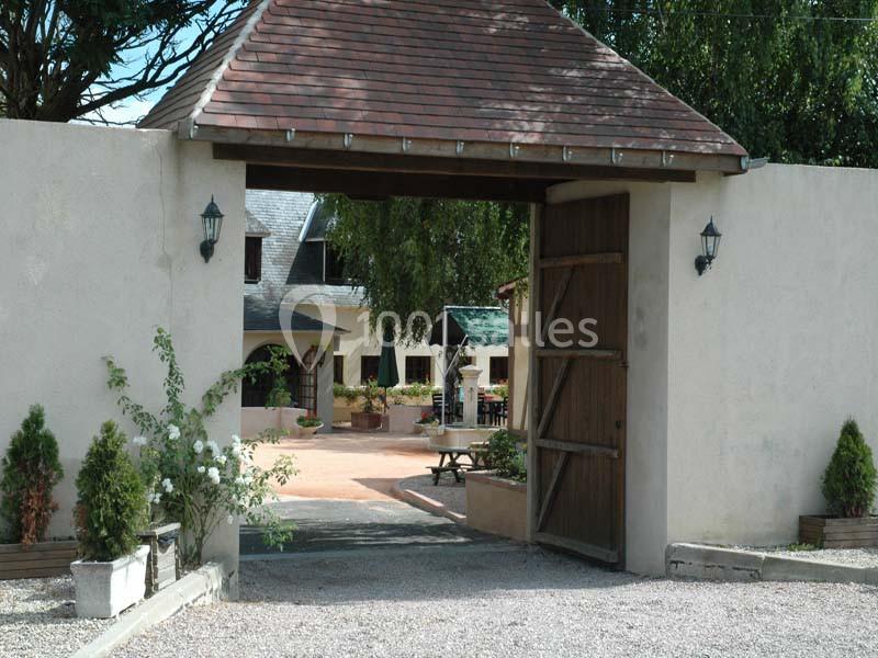Entrée d'une cour pavée avec un portail en bois ouvert, entourée de murs clairs et de végétation.