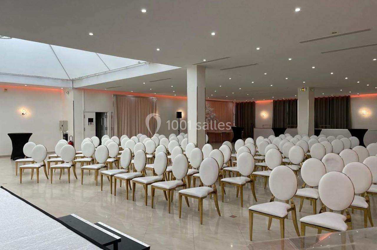 Salle lumineuse aménagée avec des rangées de chaises blanches à structure dorée, prête pour un événement.