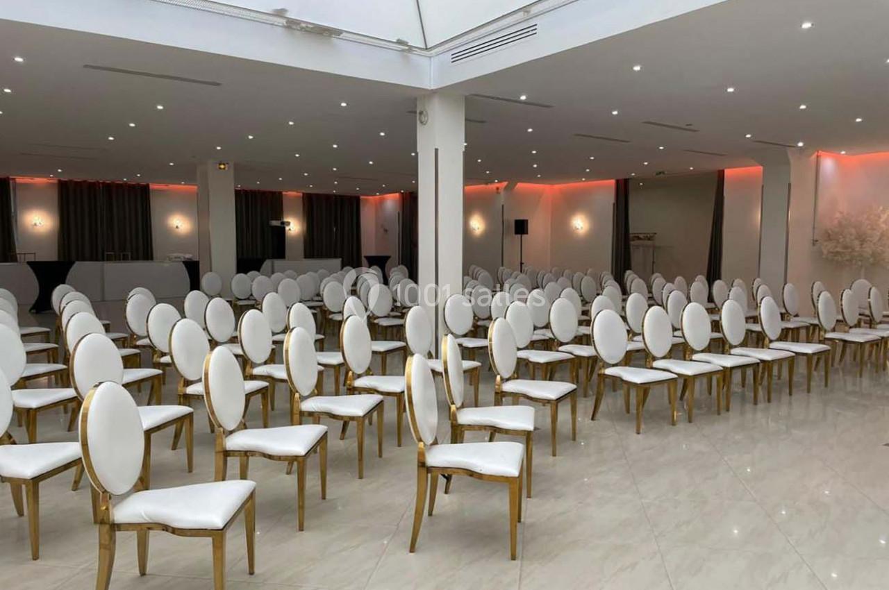 Salle de réception lumineuse avec des rangées de chaises blanches et dorées alignées sur un sol en marbre clair.