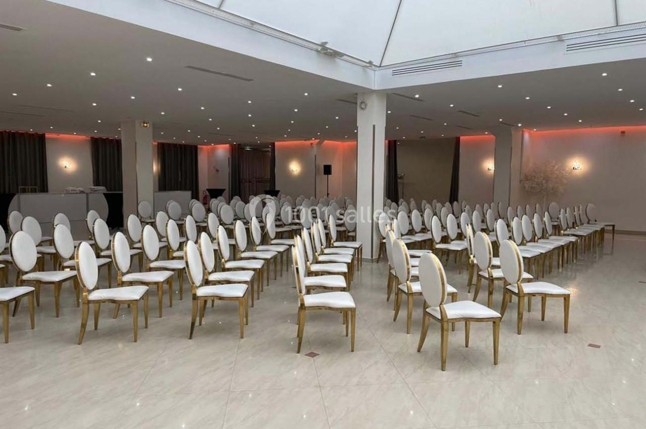 Salle de réception lumineuse avec rangées de chaises blanches et dorées alignées sur un sol en carrelage clair.
