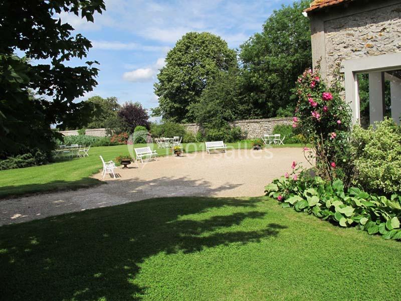 Jardin avec pelouse, bancs blancs, rosiers fleuris et arbres, entouré d'un mur en pierre sous un ciel dégagé.