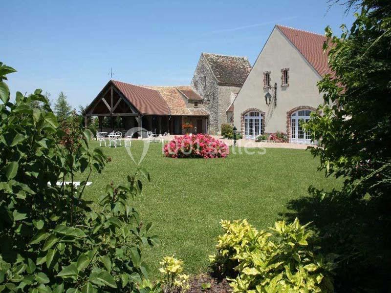 Jardin verdoyant avec pelouse, massifs fleuris et bâtiments en pierre aux toits en tuiles sous un ciel dégagé.