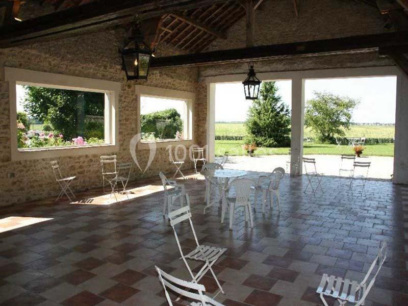 Salle ouverte avec des murs en pierre, des fenêtres donnant sur un jardin, et des chaises blanches disposées sur un sol…
