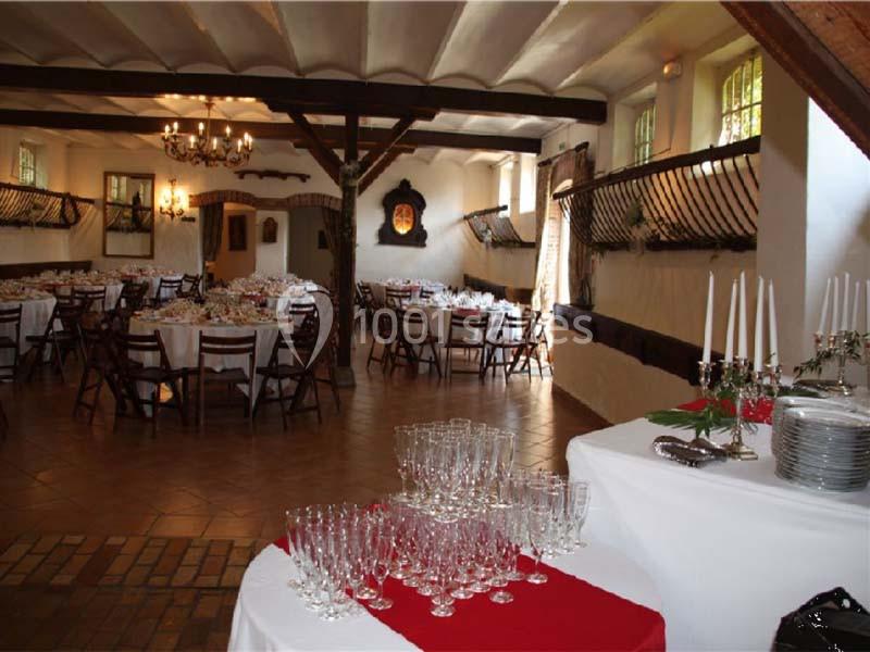 Salle de réception rustique avec tables dressées, chandeliers suspendus et verres disposés sur une table blanche.