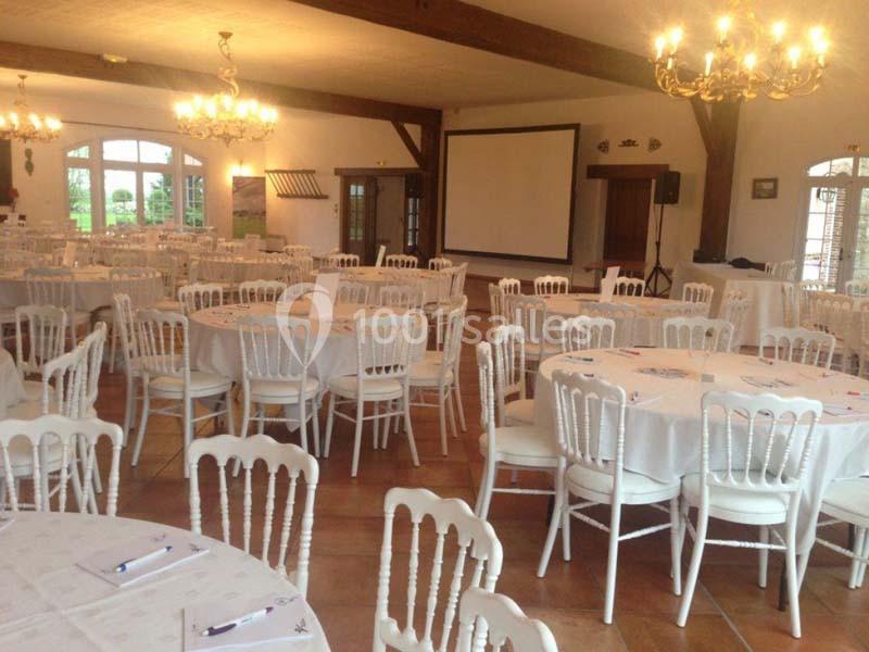 Salle de réception avec tables rondes dressées, chaises blanches et écran de projection en arrière-plan.