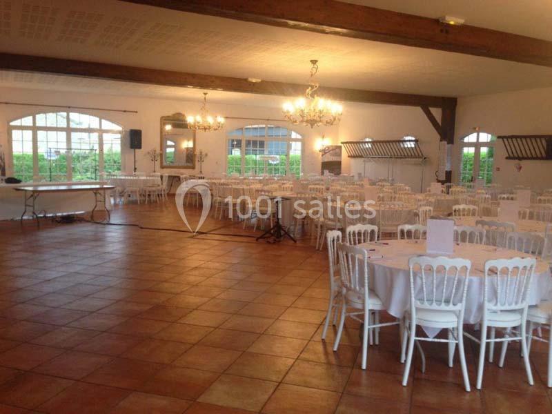 Salle de réception lumineuse avec tables rondes dressées, chaises blanches et grandes fenêtres donnant sur un jardin.