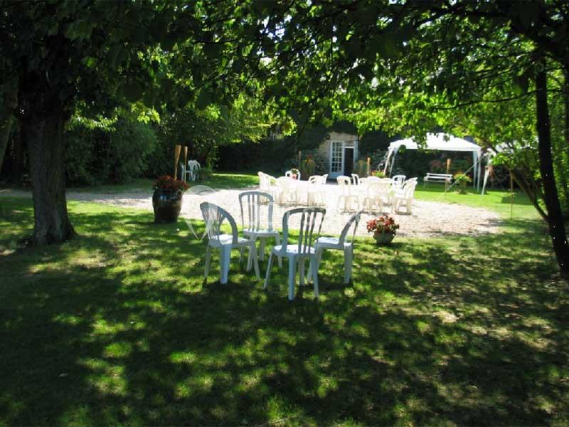 Chaises blanches en plastique disposées sur une pelouse ombragée, avec une tente et un jardin en arrière-plan.
