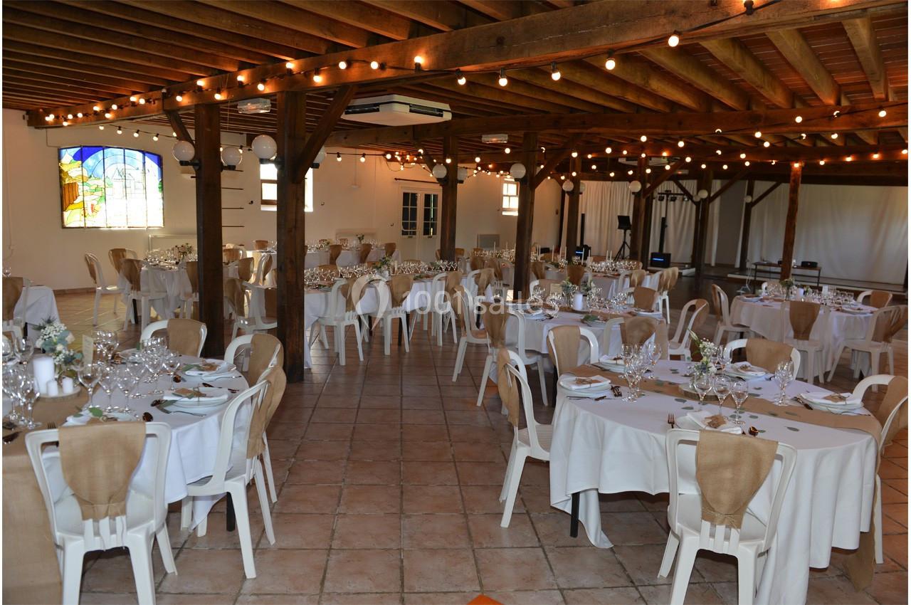 Salle de réception décorée avec des tables rondes dressées, nappes blanches et guirlandes lumineuses suspendues.