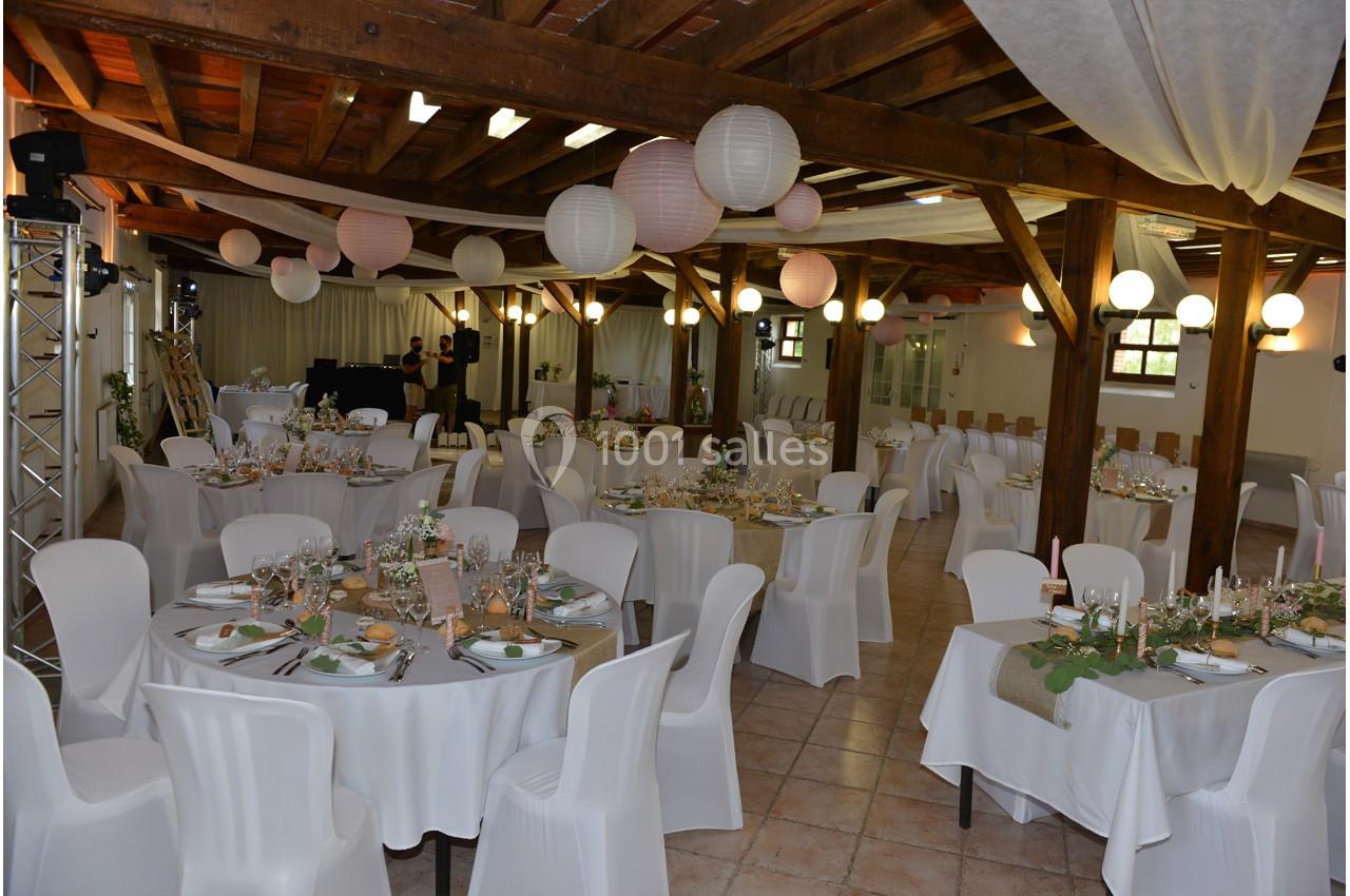 Salle de réception décorée pour un événement, avec tables rondes dressées, chaises blanches et lanternes suspendues.