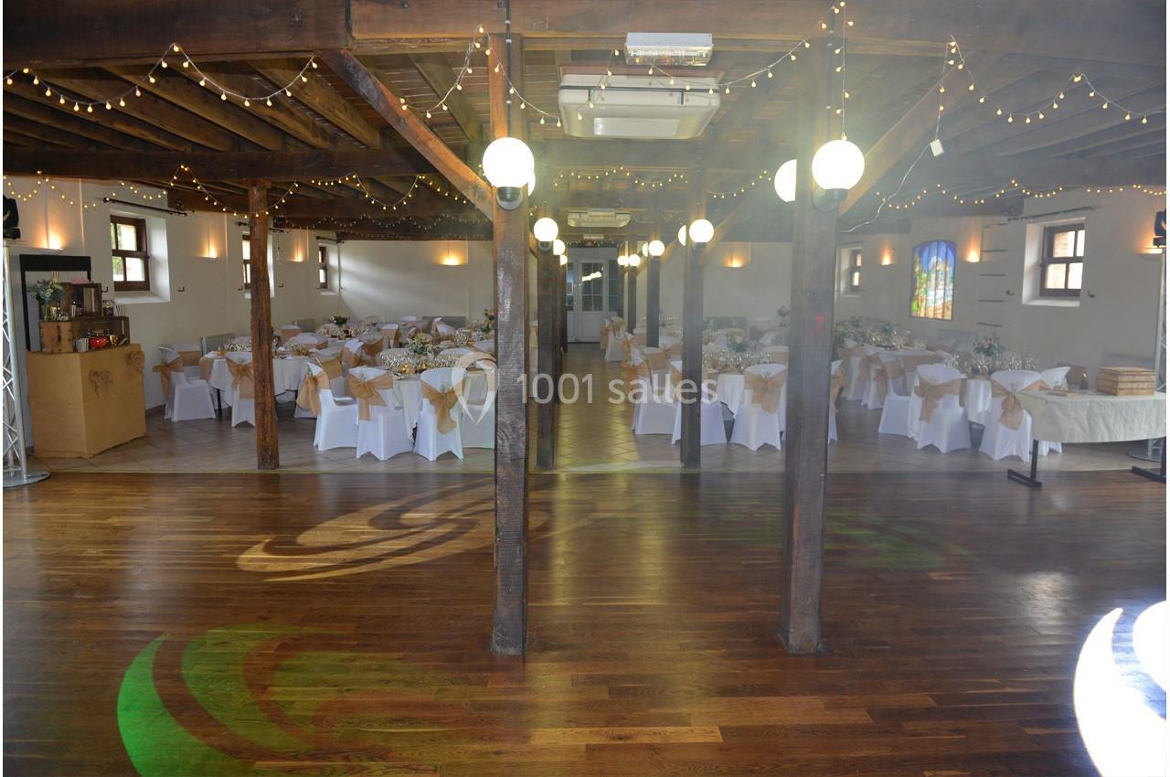 Salle de réception avec tables décorées de nappes blanches et chaises, éclairée par des guirlandes lumineuses suspendues.