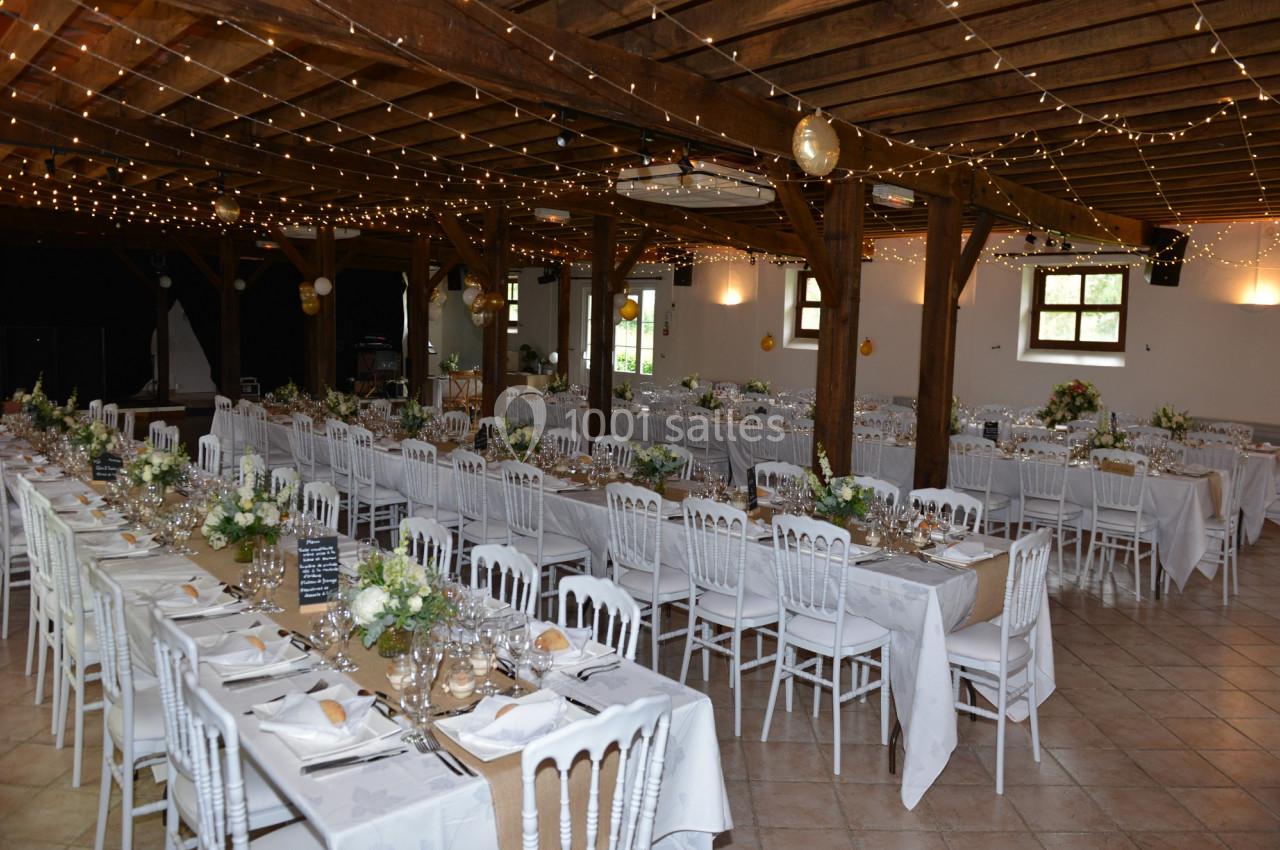 Grande salle de réception décorée avec des guirlandes lumineuses, des tables dressées et des chaises blanches.