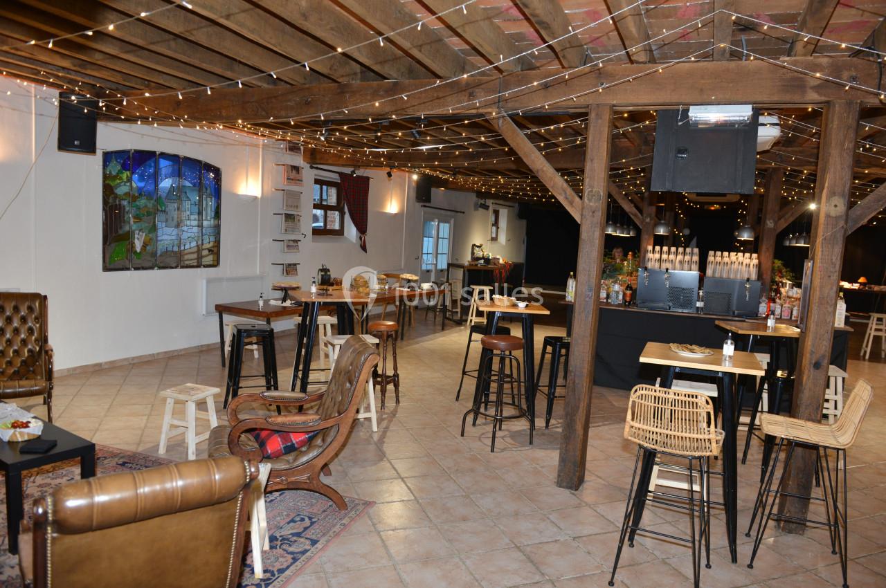Salle aménagée avec des tables hautes, des chaises en bois et un bar, éclairée par des guirlandes lumineuses suspendues.