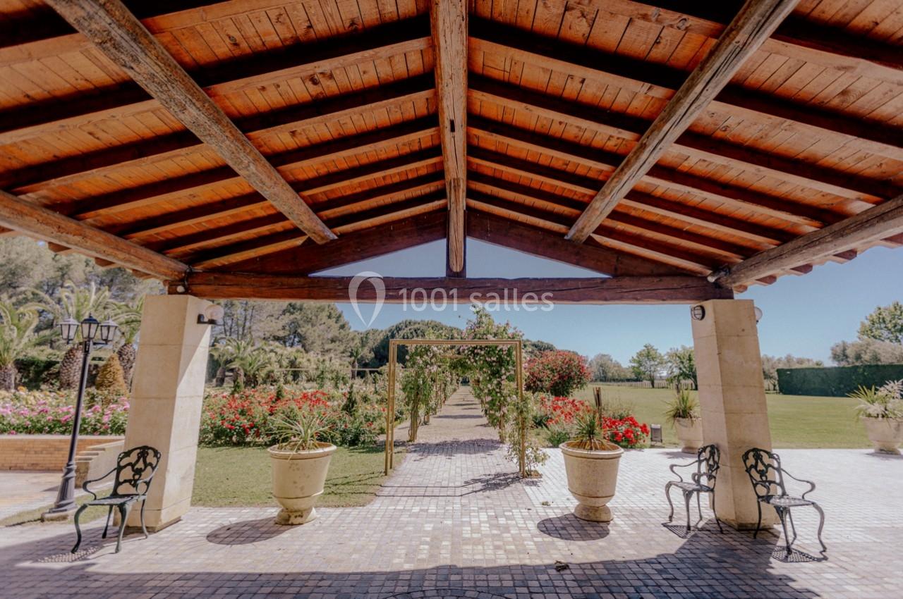Structure en bois avec toit ouvert, encadrant une allée fleurie bordée de verdure et de pots décoratifs.