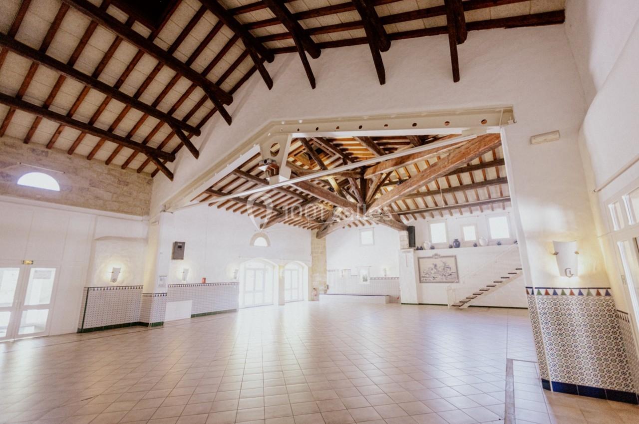 Grande salle lumineuse avec plafond en poutres apparentes, murs blancs et carrelage au sol, éclairée par des fenêtres.