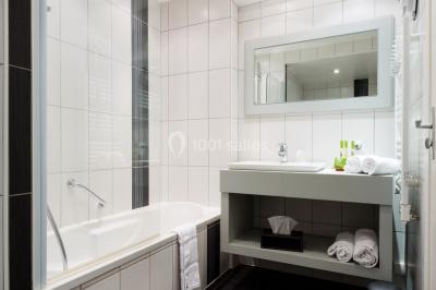 Chambre lumineuse avec lit double, fauteuils, table basse, valise et rideaux blancs laissant entrer la lumière naturelle.