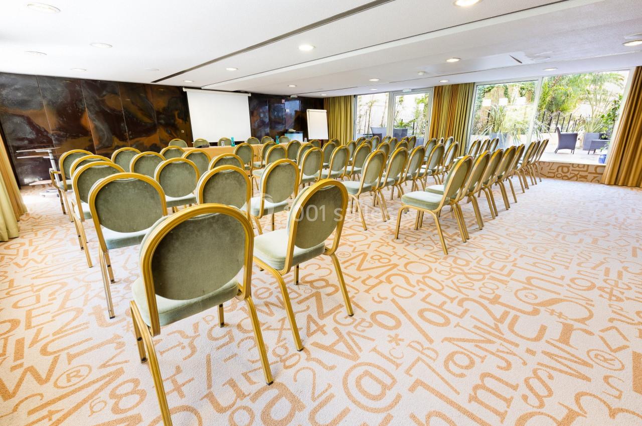 Salle de conférence lumineuse avec des rangées de chaises vertes, un écran de projection et des baies vitrées donnant sur un…
