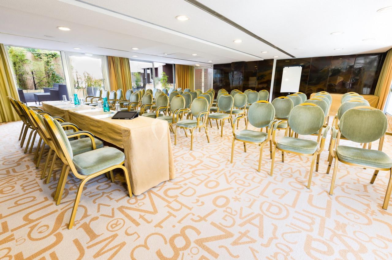 Salle de conférence lumineuse avec des rangées de chaises vertes, une table recouverte d'une nappe beige et un tableau blanc.
