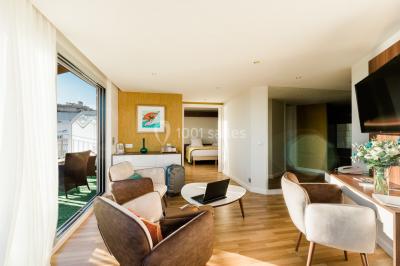 Chambre lumineuse avec lit double, fauteuils, table basse, valise et rideaux blancs laissant entrer la lumière naturelle.