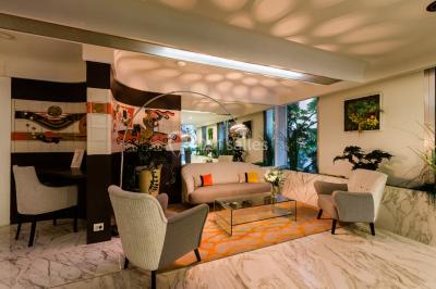 Chambre lumineuse avec lit double, fauteuils, table basse, valise et rideaux blancs laissant entrer la lumière naturelle.