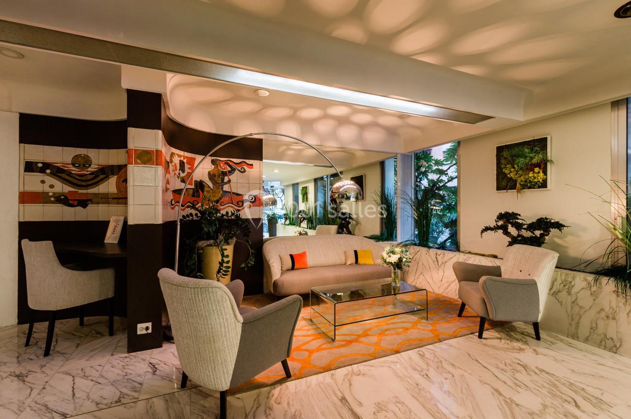 Salon moderne avec canapé, fauteuils gris, table basse en verre, décorations murales colorées et plantes vertes.