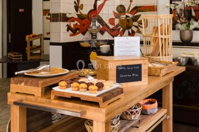 Chambre lumineuse avec lit double, fauteuils, table basse, valise et rideaux blancs laissant entrer la lumière naturelle.